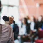 Stock photo of a woman speaking during a workplace presentation to a group of people.