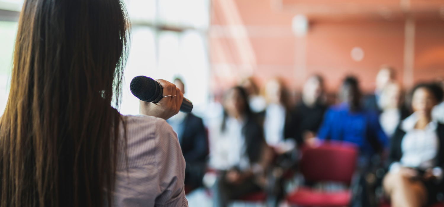 Stock photo of a woman speaking during a workplace presentation to a group of people.