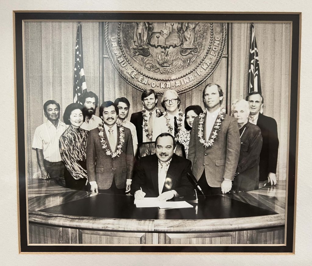 Bill signing ceremony for the UIPA. Governor John D. Waihe'e III is sitting at a desk, holding a pen, over the bill, which is on the desk.  11 people are standing behind the Governor.  Behind the group, is a large State of Hawaii seal and two flags, the US flag and Hawaiian flag.