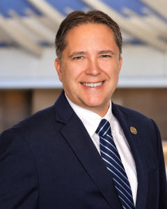 Headshot of Caucasian male in a dark business suit and tie with the Hawaii State Capitol as the background.