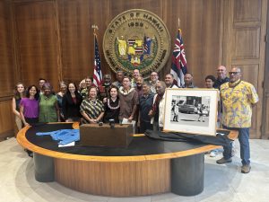 23 people stand at the Lt. Governor's desk at the exhibit of Martin Luther King Jr. photos given to the state. On one side of the desk is one of the framed photos from the 19654 Selma to Montgomery March in which six Hawaii residents took part.